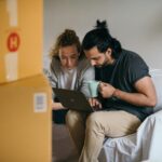 Couple surrounded by boxes checks laptop details on moving day.